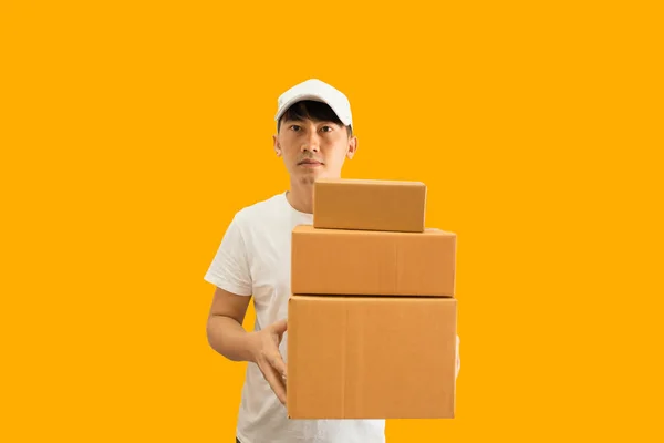 Young Asian delivery man wearing cap and white blank t-shirt holding parcel post box isolated on yellow background. express delivery service concept.