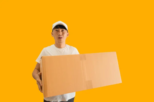 Young Asian delivery man wearing cap and white blank t-shirt holding parcel post box isolated on yellow background. express delivery service concept.