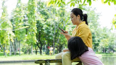 Anne ve kızı parkta oynarken birbirlerine sarılıyorlar. Asyalı anne ve çocuklar dışarıda cep telefonu kullanıyorlar..