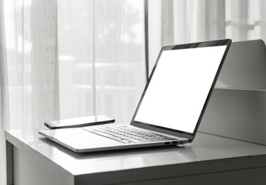 Ev içi veya ofis arka planında beyaz masadaki boş ekran ve akıllı telefona sahip bilgisayar laptopu.