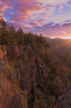 Güneşin ilk ışıkları New Mexico 'daki SantaFe Ulusal Ormanı' ndaki bir uçurumun kenarını aydınlatıyor. Mavi gökyüzü, sabah parıltısıyla parlayan alçak bulutlarla doludur. Büyük yeşil ağaçlar aşağıdaki vadiyi gözlüyor. 
