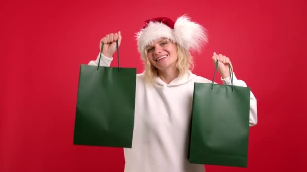 Ventas y compras navideñas. Retrato de Mujer alegre positiva en sombrero de Santa con bolsas verdes sobre fondo rojo estudio. Prepárate para el logo. Comprador adicto quieren tienda todas las gangas. — Vídeo de stock