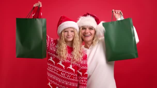 Ventas y compras navideñas. Retrato de mamá y su hija positivas en Santa sombrero con bolsas de compras verdes sobre fondo rojo estudio. Prepárate para el logo. Comprador adicto quieren tienda todas las gangas. — Vídeos de Stock