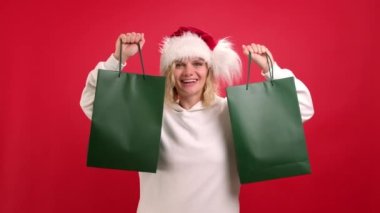 Noel satışları ve alışveriş. Kırmızı stüdyo arka planında yeşil alışveriş torbalarıyla Noel Baba şapkalı neşeli kadının portresi. Logo için hazırlan. Bağımlı müşteriler tüm pazarlıkları istiyor..