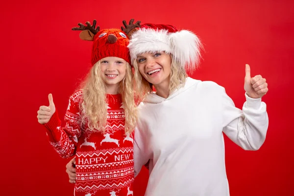 Jul semester Porträtt lycklig familj av två I Santa hattar mor och dotter stående och ler på röd studio bakgrund. visar gest tummen upp. — Stockfoto