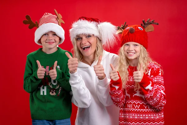 Julhelger Porträtt lycklig familj av tre I Santa hattar mor och barn stående och ler på röd studio bakgrund. visar gest tummen upp — Stockfoto