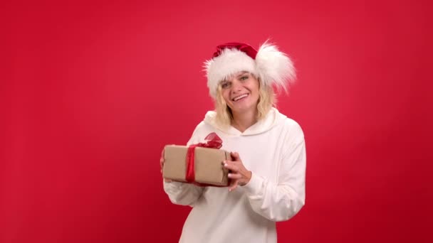 Święta Bożego Narodzenia sprzedaż i zakupy. Portret pozytywnej wesołej dziewczyny w kapeluszu Santa taniec z pudełka na czerwonym tle studio — Wideo stockowe