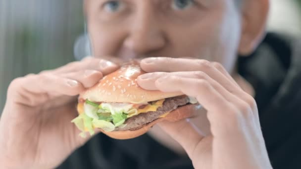 Mund aus nächster Nähe Man Student isst Burger im Fast-Food-Restaurant Juicy köstlichen großen Burger mit Fleisch, Salatkäse und Tomaten im Inneren. Ungesunde Ernährung, Snacks, Junk Food. — Stockvideo