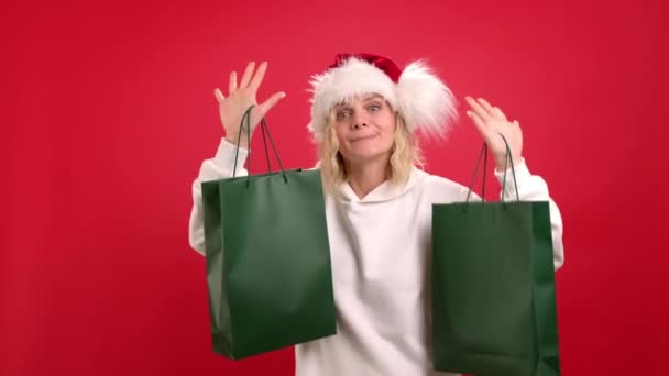 Ventas y compras navideñas. Retrato de Mujer alegre positiva en sombrero de Santa con bolsas verdes sobre fondo rojo estudio. Prepárate para el logo. Comprador adicto quieren tienda todas las gangas. — Vídeos de Stock