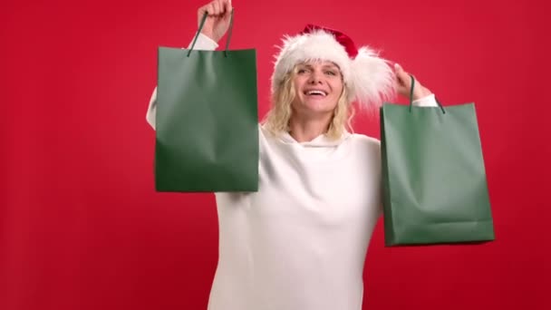 Kerstvakantie verkoop en winkelen. Portret van positieve vrolijke vrouw in Santa hoed met groene boodschappentassen op rode studio achtergrond. Maak je klaar voor het logo. Verslaafde shopper wil winkel alle koopjes. — Stockvideo