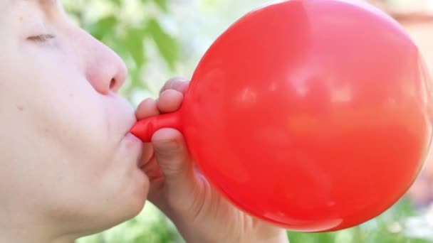 Teenager boy is blowing air balloon. Handsome redhead boy inflating red heart shaped balloon holding balloon in hands. Mouth closeup. Happy birthday party, celebration, valentines day — Stock Video