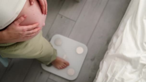 Молодая женщина измеряет свой беременный вес стоя на весах. Подготовка к родам, Девочка с большим животом развилась здоровой беременностью — стоковое видео