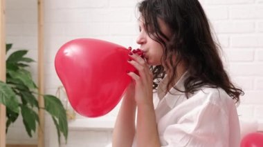 Kız evde balon şişiriyor. Yakışıklı esmer kız. Kırmızı kalp şeklinde balon şişiriyor. Sevgililer günü partisi, kutlama, flört ve sürpriz hazırlıkları.