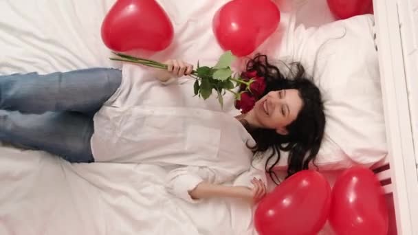 Ragazza eccitata che cade sul letto palloncini a forma di cuore rosso con mazzo di fiori. La donna gioisce del dono il giorno di San Valentino. Ragazza rilassata con i capelli lunghi che esprimono emozioni positive nel giorno di San Valentino — Video Stock