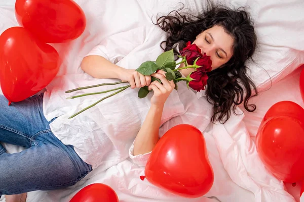 Podekscytowana dziewczyna leżąca na łóżku czerwony kształt serca balony z bukietem kwiatów Kobieta raduje się prezent na Walentynki. Łatwa dziewczyna z długimi włosami wyrażająca pozytywne emocje w Walentynki Zdjęcia Stockowe bez tantiem