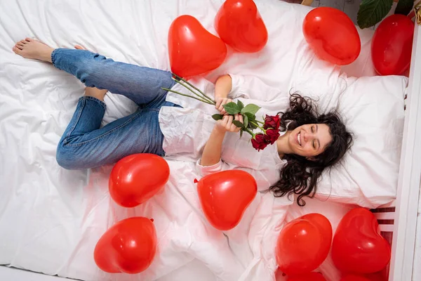 Podekscytowana dziewczyna leżąca na łóżku czerwony kształt serca balony z bukietem kwiatów Kobieta raduje się prezent na Walentynki. Łatwa dziewczyna z długimi włosami wyrażająca pozytywne emocje w Walentynki Obrazek Stockowy