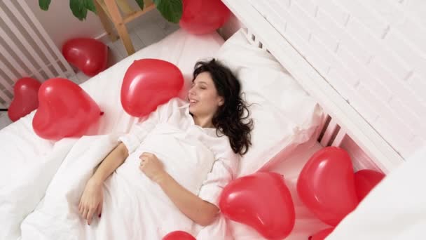 Європейська дівчина Брюнет прокидається на ліжку з кульками червоної серцевої форми. Ранок любові подарунок у день примхи — стокове відео