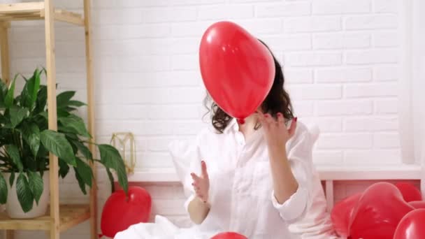 Brünettes europäisches Mädchen freut sich, auf dem Bett mit roten herzförmigen Luftballons zu spielen. Morgen Liebe Überraschungsgeschenk am Valentinstag — Stockvideo