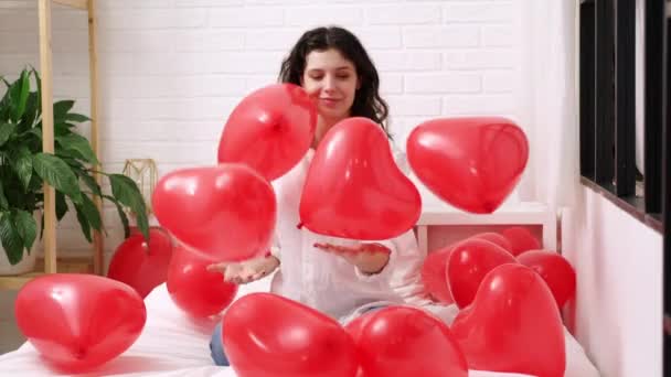 Brünettes europäisches Mädchen freut sich, auf dem Bett mit roten herzförmigen Luftballons zu spielen. Morgen Liebe Überraschungsgeschenk am Valentinstag — Stockvideo