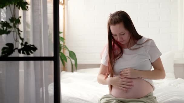 Беременная будущая мать ласкает живот. Женщина руки трогают ее животик, ожидая ребенка, сидя на кровати Подготовка к родам, девушка большой живот продвинутой беременности — стоковое видео