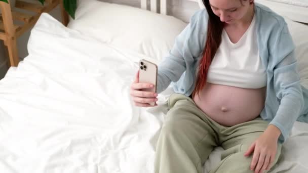 Красивая веселая беременная женщина делает селфи фото с помощью мобильного телефона, сидя на кровати дома. беременность с большим животом. — стоковое видео