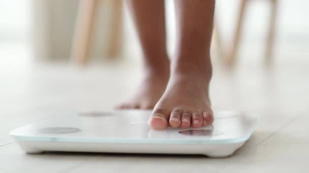 Fekete lány lábai állnak a mérlegen, hogy megmérjék a súlyt. Afrikai-amerikai Nő mezítláb, súlyskálával otthon. fogyókúra, ellenőrzés és mérés. — Stock videók