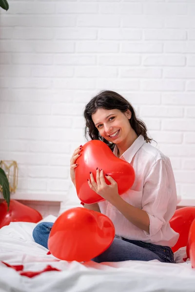 Портрет взволнованной Брюнетка Европейская девушка радуется, играя на кровати с красными шарами формы сердца. Утренний подарок на день Святого Валентина — стоковое фото