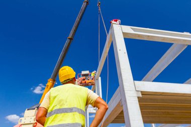 Güvenlik yeleği ve sarı miğferi olan inşaat işçisinin arkasından, inşaat için beton kirişi yöneten mobil vinç görüntüsü.