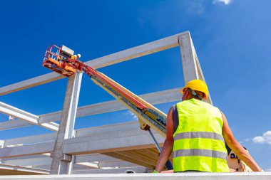 Güvenlik yeleği ve sarı miğferi olan inşaat işçisinin arkasından, inşaat için beton kirişi yöneten mobil vinç görüntüsü.