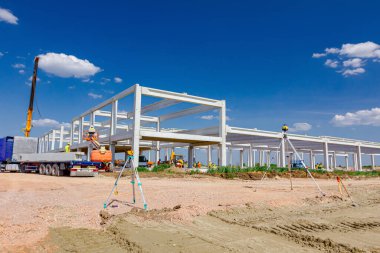 Mobil vinç endüstriyel binanın beton iskeletini monte ediyor, makinelerle, insanlar çalışıyor. İnşaat alanında lazer seviyelendirme diğer cihazlara sahip toplam merkez aygıt.