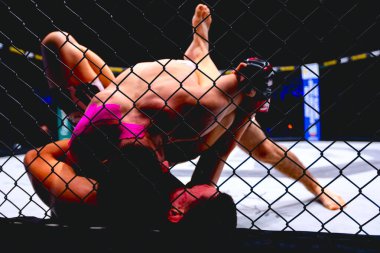 İki güçlü kaslı profesyonel MMA dövüşçüsü, birbirlerine kenetlenmişler, sekizgen katta parterre güreşiyorlar..