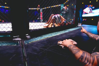 Dövmeli antrenör eli havluyla, dövüşçüleri destekliyor, sekizgen katta ringde dövüşüyorlar..