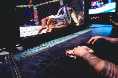 Dövmeli antrenör eli havluyla, dövüşçüleri destekliyor, sekizgen katta ringde dövüşüyorlar..