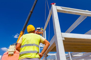 Güvenlik yeleği ve sarı miğferi olan inşaat işçisinin arkasından, inşaat için beton kirişi yöneten mobil vinç görüntüsü.