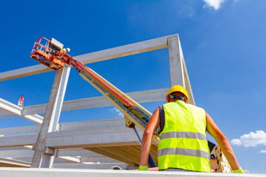 Güvenlik yeleği ve sarı miğferi olan inşaat işçisinin arkasından, inşaat için beton kirişi yöneten mobil vinç görüntüsü.