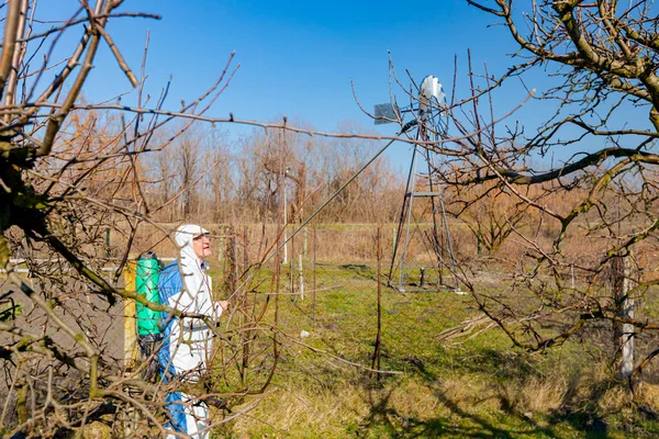 保護服の農家は 春の初めに真菌病や害虫から化学物質でそれらを保護するために長い噴霧器を使用して果樹園で果物の木を噴霧 — ストック写真