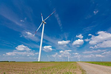 Tozlu yol, tarımsal alanlarda duran ve temiz yenilenebilir elektrik enerjisi üreten pek çok büyük rüzgar türbini arasında ilerliyor..