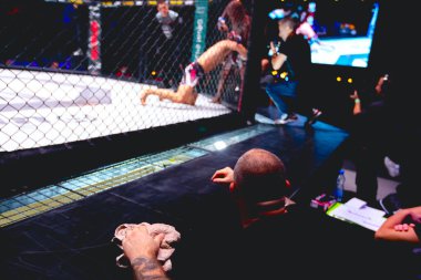 Dövmeli antrenör eli havluyla, dövüşçüleri destekliyor, sekizgen katta ringde dövüşüyorlar..