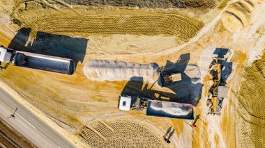 Üst tarafta, kargoyu, kesiri, küçük ezilmiş taşı boşaltmak için karavanlarını kaldıran iki kamyonun tepesinde buldozer sürüngeninin ittiği, inşaat sahasında dengelediği küçük bir taş var..