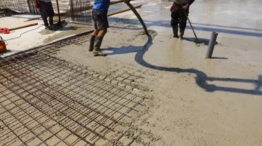 Construction worker is directing the pump tube on right direction, pouring layer of concrete in building foundation. Riggers, workers are using rake to leveling concrete covering square reinforcement.