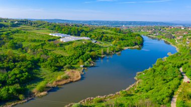 Above view on lake, forest, colorful hilly landscape with cottage settlement, several greenhouses on cultivated arable plots, farmland, city in the distance.