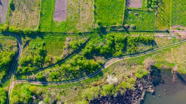 Above top view, overhead, on the end of the lake surrounded by green forest and several cultivated plots with cottages.