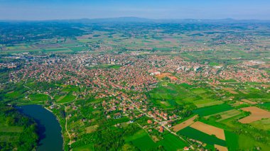 Above view on lake, forest, colorful hilly landscape with cottage settlement, several greenhouses on cultivated arable plots, farmland, city in the distance.