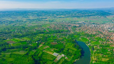 Above view on lake, forest, colorful hilly landscape with cottage settlement, several greenhouses on cultivated arable plots, farmland, city in the distance.