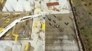 Aerial view, dolly move and orbit lateral, construction worker is directing the pump tube as pouring layer of concrete in building foundation. Riggers, workers are leveling concrete over reinforcement.