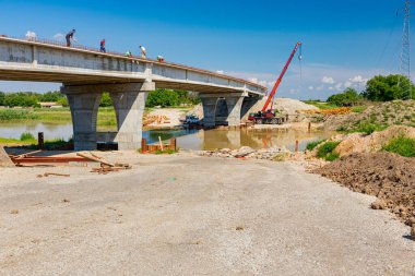 Team of workers are working on the unfinished bridge, road over river is under construction, building site.