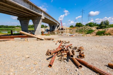 İnşaat alanında yere yerleştirilmiş paslanma borularından oluşan yığınla parçalanmış çelik yapı iskelesine bakın. Köprü arka planda..