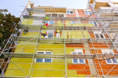 View from below on fenced thermally insulated building. Riggers on scaffolding are applying thermal isolation rock wool on the unfinished brick wall of residential build under construction.