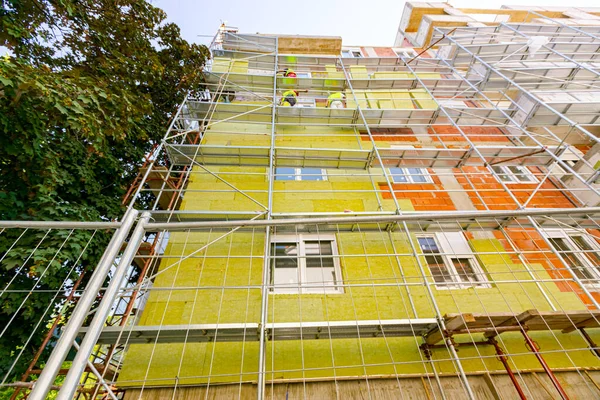 View from below on fenced thermally insulated building. Riggers on scaffolding are applying thermal isolation rock wool on the unfinished brick wall of residential build.