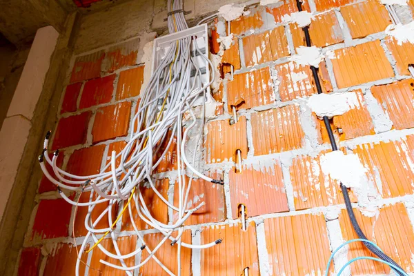 Bundle of various cables hang from a plastic fuse box mounted on the wall, building under construction, work in progress. Installation of electrical wires and cables.